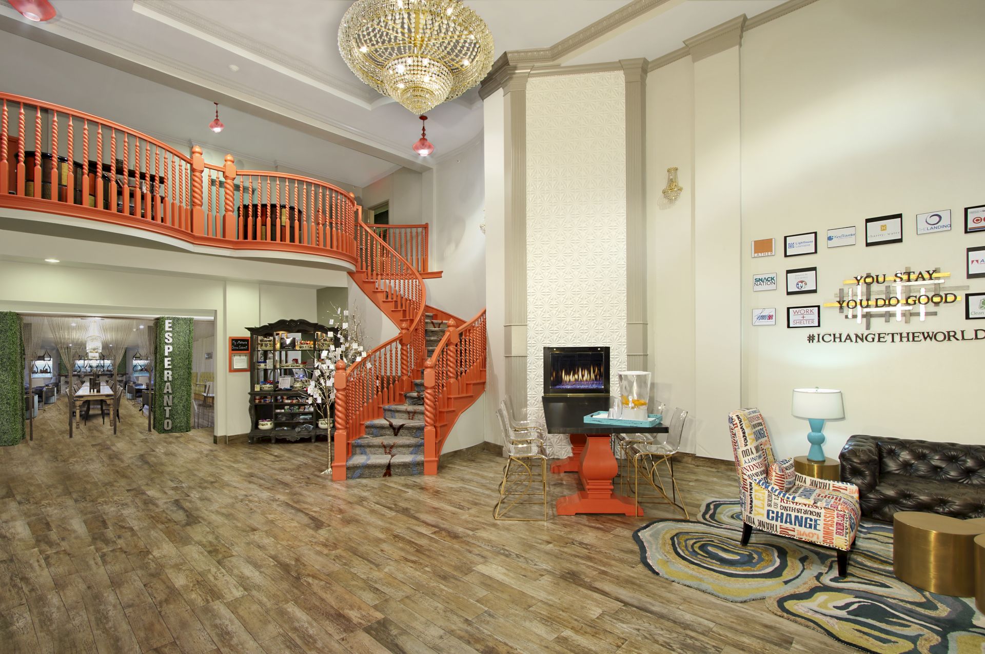A spacious room with a spiral staircase, chandelier, seating, wall decor, and flooring. A sign reads "YOU STAY, TOGETHER WE #CHANGETHEWORLD" ending the sentence.