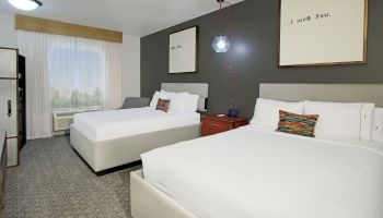 The image shows a hotel room with two double beds, modern decor, a nightstand with a lamp, and artwork on the walls.