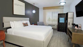 A modern hotel room with a queen bed, a nightstand, a TV on the wall, a desk, a wardrobe, and a chair by the window with natural light flowing in.