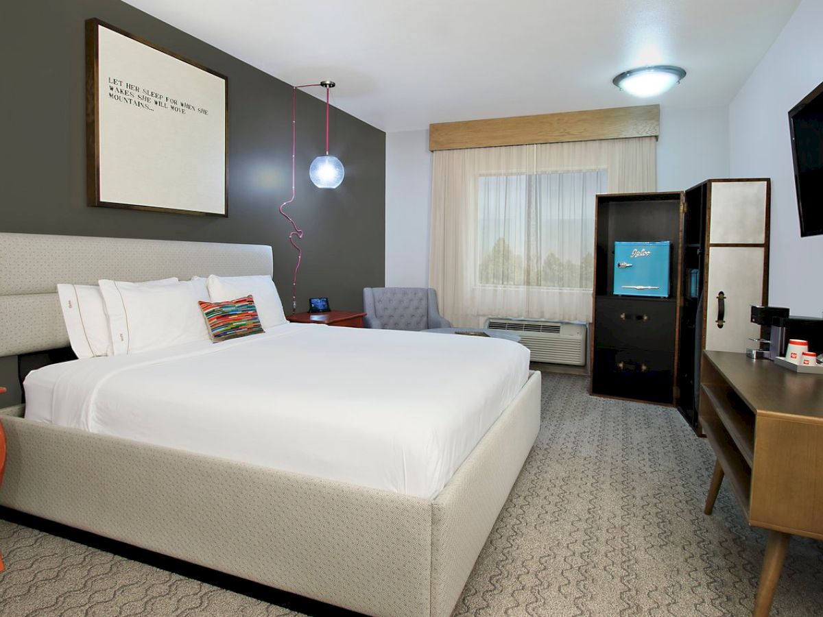 The image shows a modern hotel room with a neatly made bed, large window, armchair, wall art, TV, desk, and a mini-fridge.