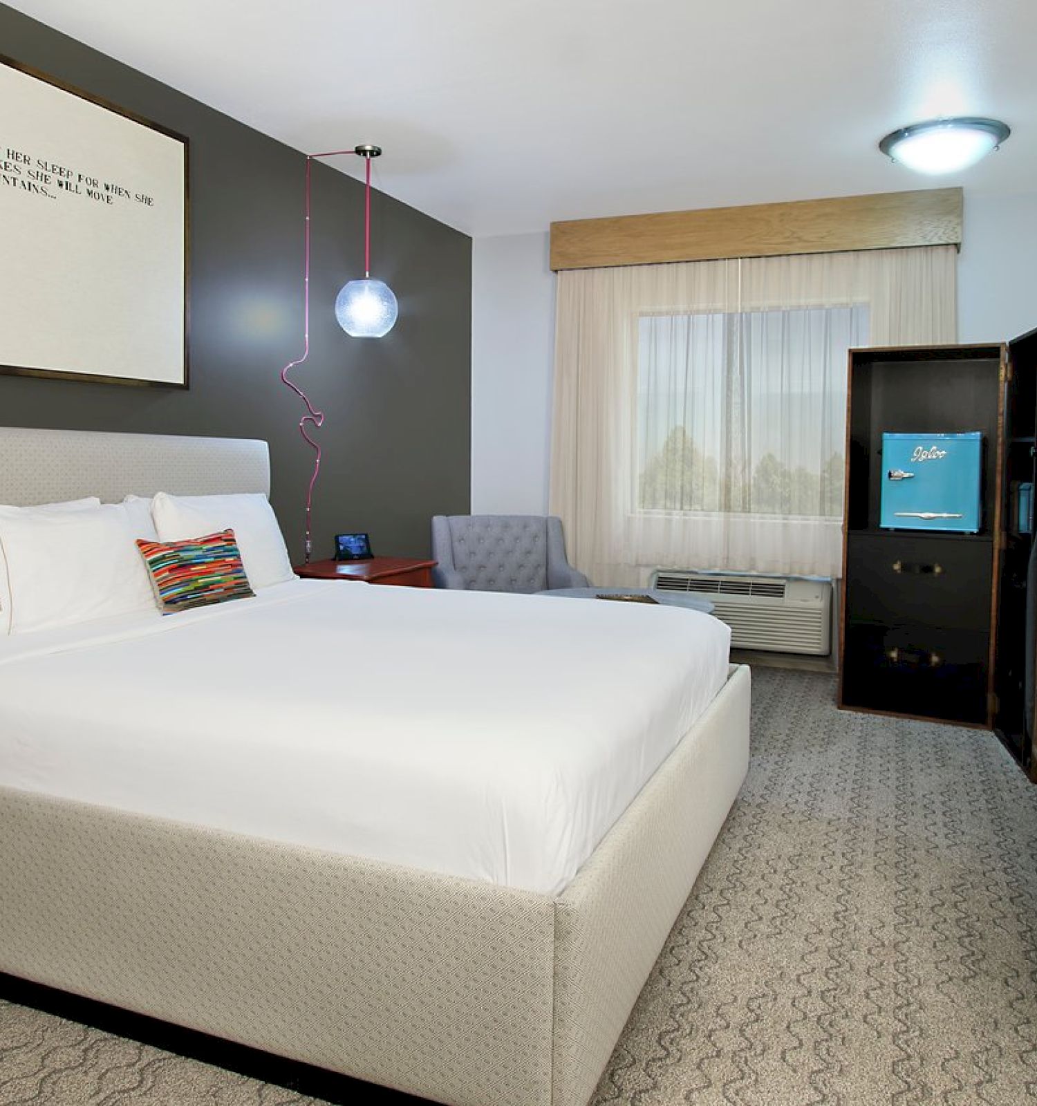 The image shows a modern hotel room with a neatly made bed, large window, armchair, wall art, TV, desk, and a mini-fridge.