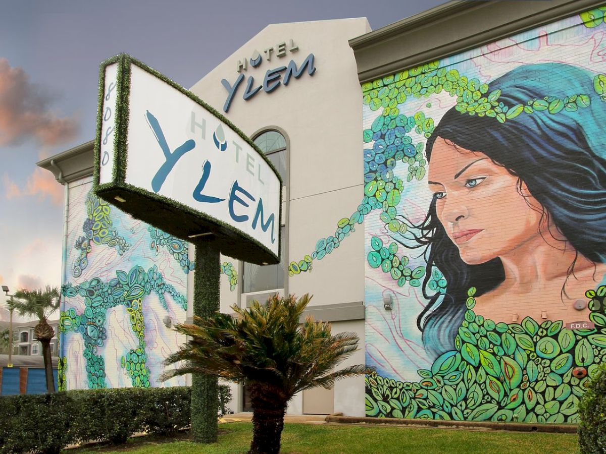 The image shows the exterior of Hotel Ylem with a large mural of a woman surrounded by leaves, and prominent signage displaying the hotel's name.