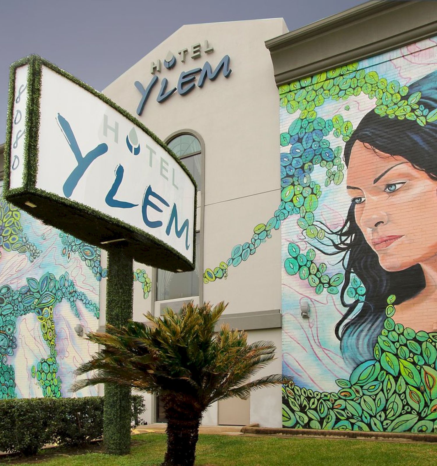 The image shows the exterior of Hotel Ylem with a large mural of a woman surrounded by leaves, and prominent signage displaying the hotel's name.