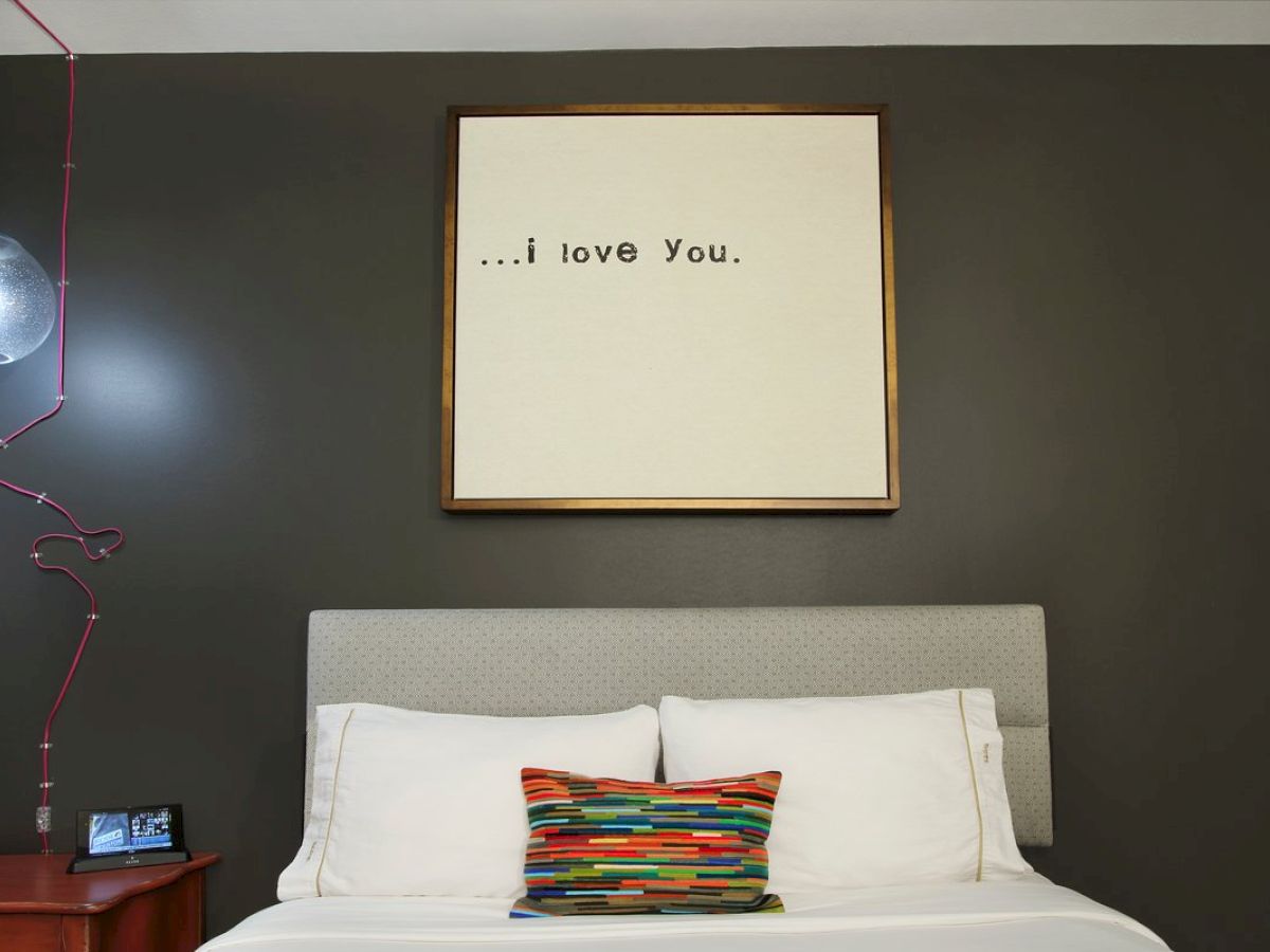 A bedroom with a dark gray wall, a bed with white pillows and a colorful cushion, a side table, and wall art reading “...i love You.”.