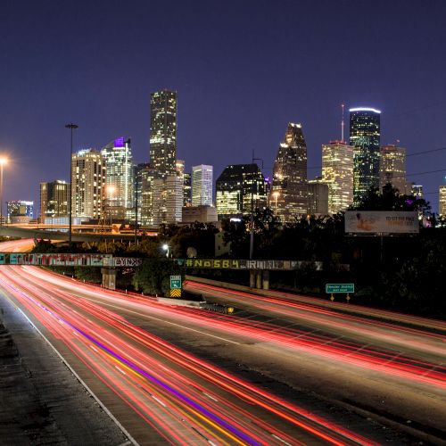 A nighttime cityscape with tall buildings illuminated, highways with light trails from moving vehicles, and clear skies.