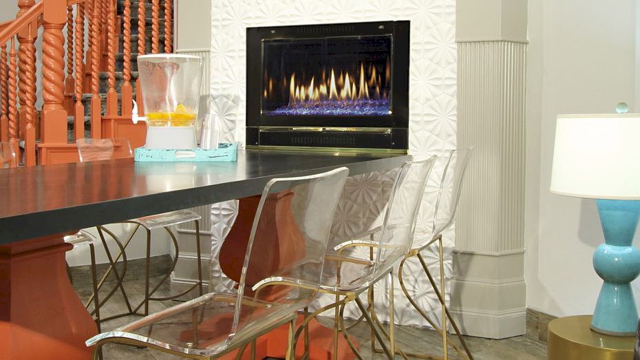 A modern room with a black table, transparent chairs, a blue lamp, and an electric fireplace, featuring orange stairs in the background.