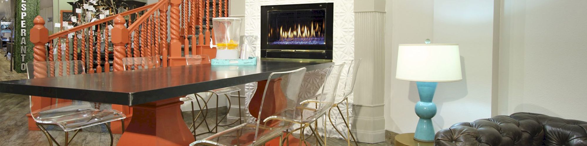 A modern room with a black table, transparent chairs, a blue lamp, and an electric fireplace, featuring orange stairs in the background.