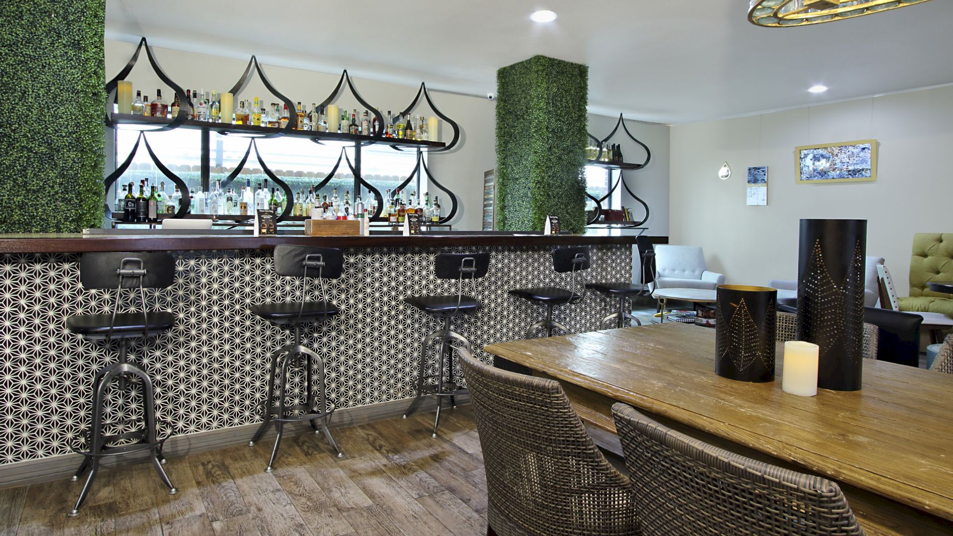 A modern bar with stylish decor, five bar stools, and a patterned tile counter. A wooden table with wicker chairs and decorative items in the foreground.