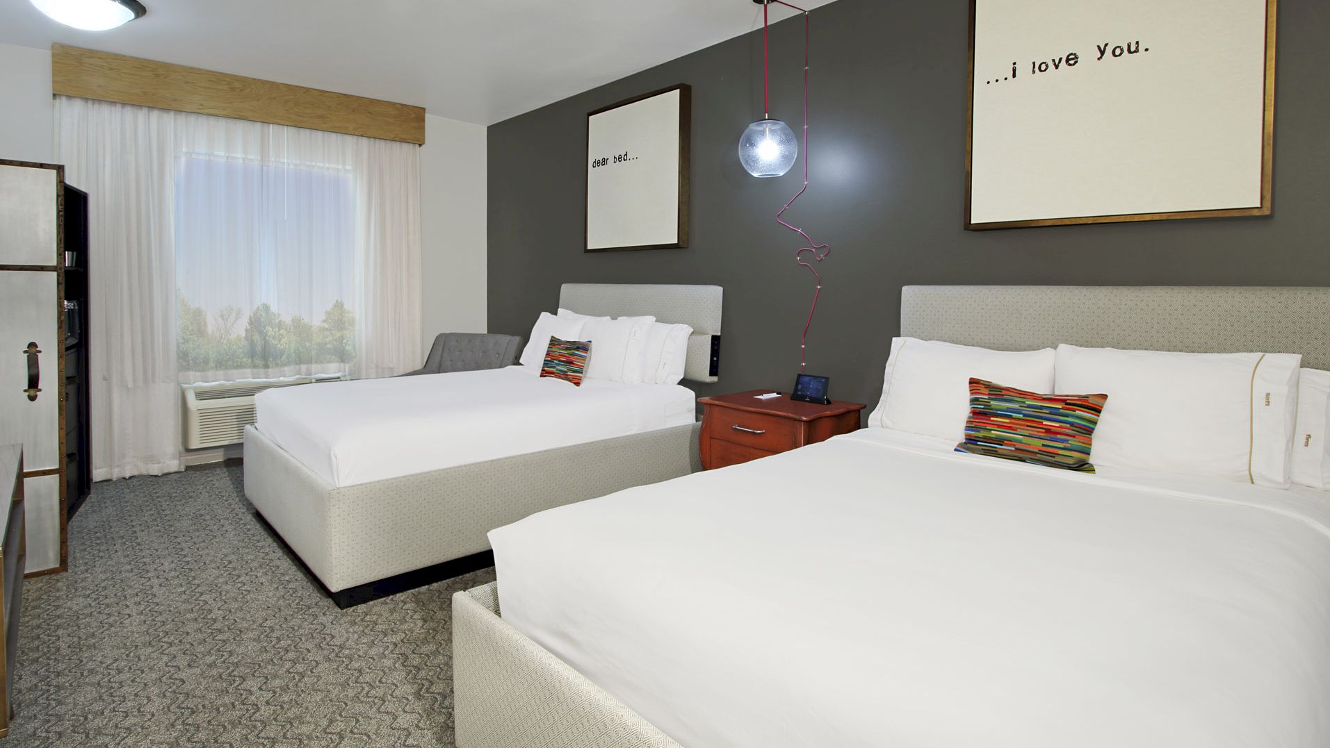 The image shows a hotel room with two double beds, a window with curtains, framed art on the wall, and pillows on the beds.