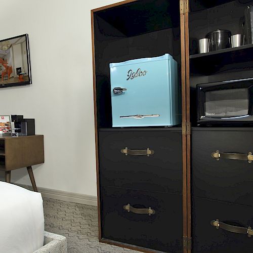 The image shows a hotel room with a blue mini fridge, microwave, coffee maker, and a TV on the wall. There is a bed visible in the foreground.