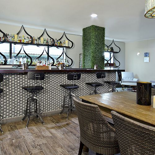 The image shows a modern bar area with high chairs, patterned tiles, and a shelf stocked with bottles. A wooden table with chairs is in the foreground.