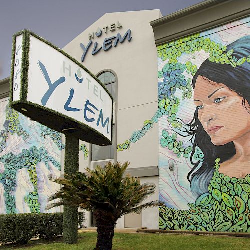 The image shows the exterior of Hotel Ylem with a large mural of a woman surrounded by greenery, and a sign also displaying 