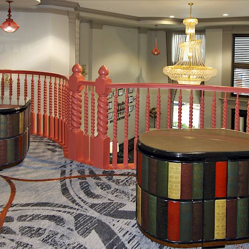 The image shows an indoor balcony with red railings, two book-themed tables, patterned carpet, and a chandelier in the background.