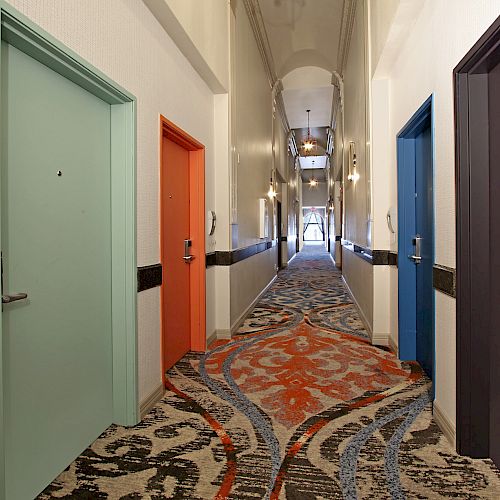 A hallway with four colorful doors (mint green, orange, blue, and dark purple) and a patterned carpet, leading to a bright area in the distance.