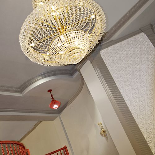 The image shows a large, ornate chandelier hanging from the ceiling, with a red staircase visible in the background and wall-mounted light fixtures.