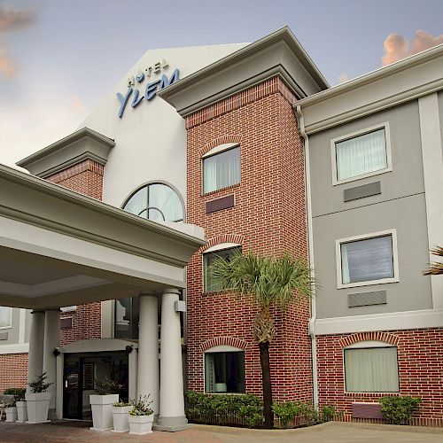 This image shows the exterior of a mid-sized hotel building with a brick facade, palm trees, and a covered entrance area.