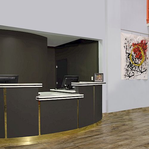 The image shows a modern reception area with a dark-colored desk, two monitors, abstract artwork on the wall, and a banner stand with information.