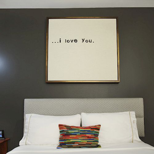 A neatly made bed with white linens and a colorful pillow; above it is a framed sign saying 