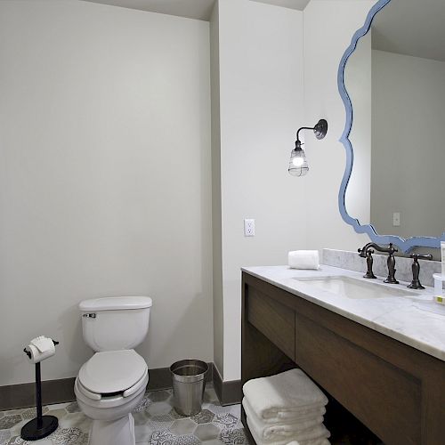 This image shows a bathroom with a toilet, a shower with a white curtain, a vanity with a blue-framed mirror above it, and various toiletries.