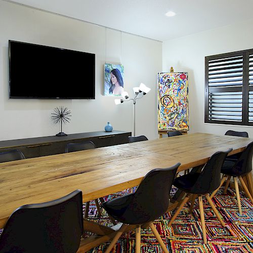 A meeting room with a large wooden table, black chairs, a TV, colorful rug, and wall art. Window with blinds and modern decorations.