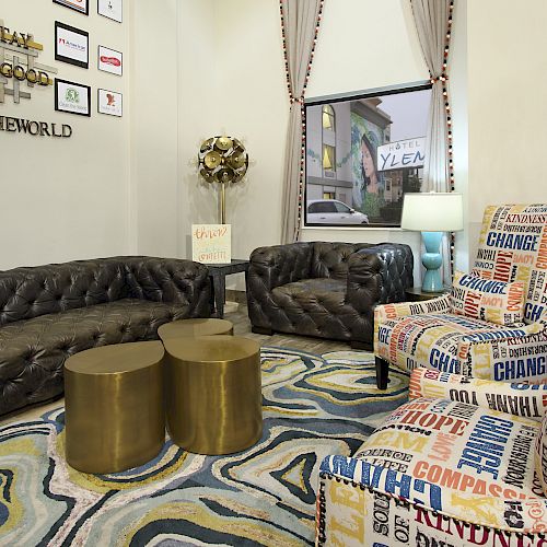 A cozy room with tufted sofas, colorful word-patterned chairs, gold round tables, and motivational wall decor. A lamp and window are also present.