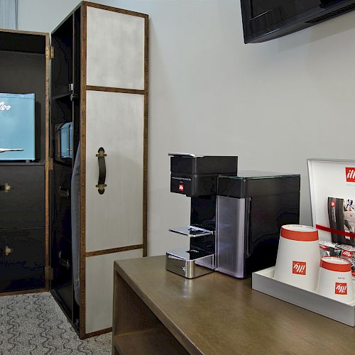 The image shows a mini fridge, a coffee maker, a set of coffee cups, and coffee supplies on a desk, likely in a hotel room.