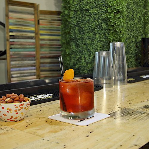 A cocktail garnished with an orange slice and a bowl of nuts are on a bar counter, with additional bar equipment in the background.