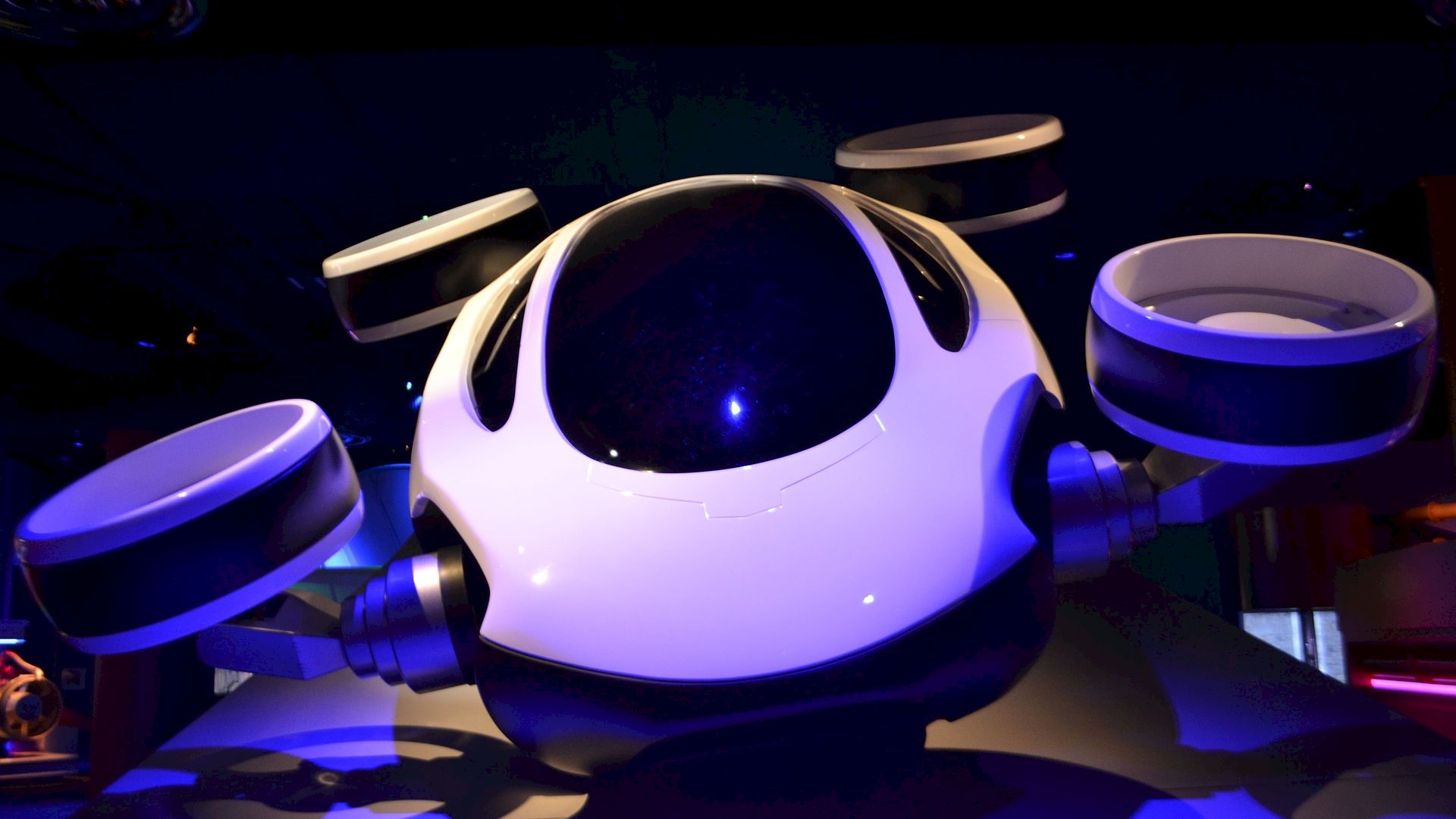 The image shows a futuristic, white and black, drone-like vehicle with four circular propellers positioned around its body, displayed in a dimly lit setting.