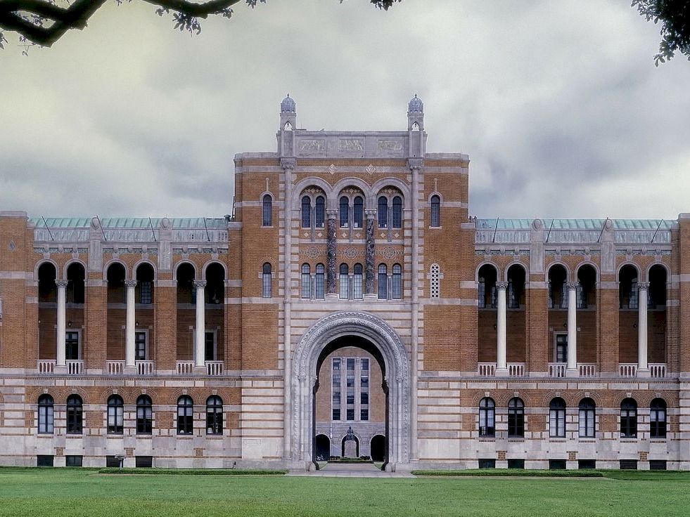 A large brick building with an arched doorway and columns, set against a cloudy sky.